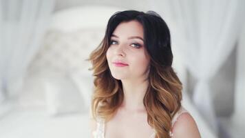 Portrait of a beautiful young bride with wavy hairdo smiling into camera. Glamour model with stylish makeup is sitting on bed and posing in the studio. video