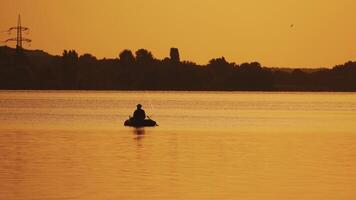 Boat with a fisherman in the sunset light floats slowly on the lake. Silhouette of a man sitting in a boat and fishing on the orange background of beautiful sunset in the river. video