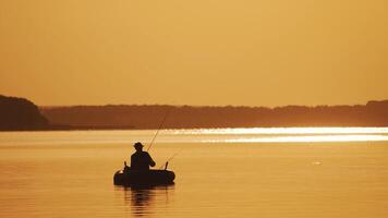 Silhouette of a man in hat fishing from the boat on the evening river background. Beautiful sunset and a fisherman catching fish from the inflatable boat in the open water. video
