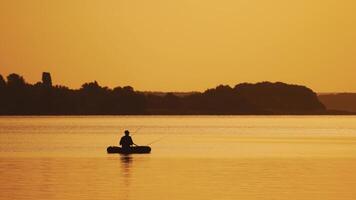 Amazing view of the evening sunset and a man in a boat fishing. Silhouette of a fisherman sitting in a row boat and catching fish on the golden evening background over the river surface. video