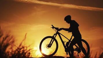 Young man with helmet walking with his bicycle on the orange sky background in the evening. Amazing view of a golden setting of a sun and a silhouette of a cyclist going next to his bike. video
