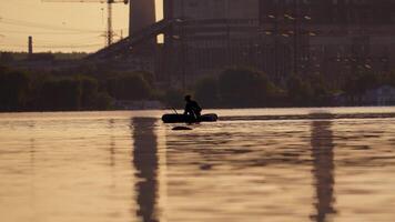 Fisherman in a boat with fishing rods in the evening. Man in hat floating in a boat and fishing on the background of a factory in the city. video