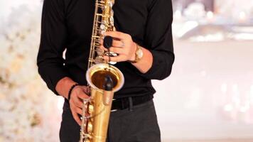 Saxophonist plays on golden saxophone indoors. A young man in black clothes is playing on a modern sax with microphone at the party. Jazz music. Close-up video