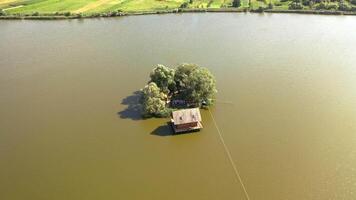 Small island with green trees and a house in the river. Aerial view of island and wooden house in the middle of a lake surrounded by water at daytime in summer. Camera moves down. video
