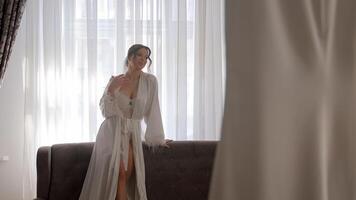 Bride posing by the window in a white robe, A bride in a delicate white robe stands by the window, looking out thoughtfully, as natural light fills the room video