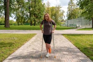 Crutches supporting her,teenage girl with broken leg in cast moves through summer park,bright, nurturing environment emphasizing her resilience and ongoing process of recovery photo