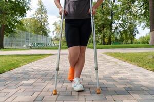 Unidentified female figure stands in parkland setting, broken limb supported by crutches - her featureless face and reserved stance present an impersonal, detached impression photo