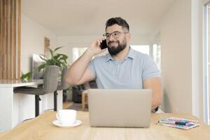 A smiling business man talking on his phone and using a laptop while working at the home office. photo