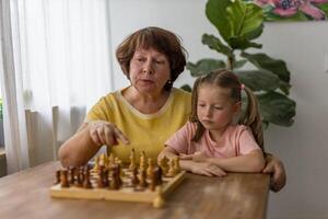 Smiling little girl plays chess with her grandmother at the table,enjoying loving and playful moment photo