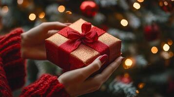 A woman is holding a red box with a gold bow on it. The box is wrapped in red and gold paper. The woman is wearing a red sweater. Concept of warmth and joy photo
