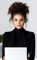 A woman with curly hair and glasses is sitting at a desk with a laptop. She is wearing a black suit and she is working photo