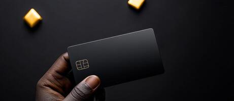 A person is holding a black credit card. The card is rectangular and has a gold border. The image has a dark background and a few yellow candies in the background photo