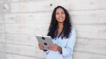 A young woman is reading on her tablet while sitting against a concrete wall in a casual setting video