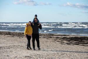 A happy family strolls along a chilly autumn beach, with their young son joyfully running ahead, creating lasting memories as the crisp wind and soothing waves embrace them in a moment of pure photo