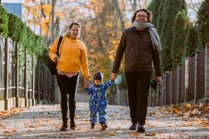 A loving couple strolls through a sunlit park with their young son, surrounded by the vibrant colors of autumn, enjoying a joyful and peaceful family moment together. photo