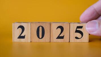 A close-up of a hand adjusting wooden blocks with black numbers to form 2025 against a vibrant yellow background video