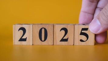 A close-up of a hand adjusting wooden blocks with black numbers to form 2025 against a vibrant yellow background video
