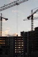 Cranes operate at a construction site during sunset, with workers visible on the building's framework as daylight fades photo