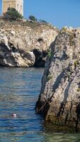A swimmer explores calm waters by a rugged shoreline, with a historical tower visible above the cliffs under clear skies photo