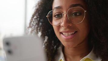 Close up face African American woman student girl female user in glasses using mobile phone app smartphone chatting browsing gadget addict smiling happy internet online searching indoors university video
