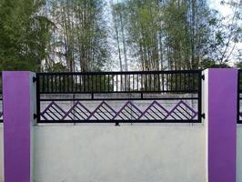 A modern fence with a purple accent wall and pillars. The fence features a geometric design and is situated in front of a bamboo grove. photo