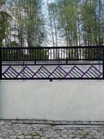 A modern fence with a purple accent wall and pillars. The fence features a geometric design and is situated in front of a bamboo grove. photo