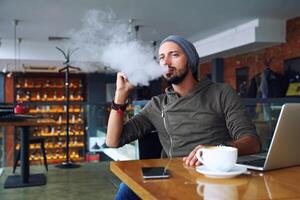 Young handsome hipster man with beard sitting in cafe with a cup of coffee, vaping and releases a cloud of vapor. Working at laptop and having a little break. With copy space. photo
