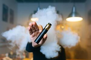 Young beard man show vaping device on his outstretched hand through a cloud of steam. Selective focus. Vaping concept. Copy space. photo