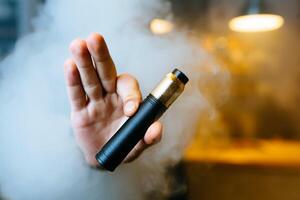 Young beard man show vaping device on his outstretched hand through a cloud of steam. Selective focus. Vaping concept. Copy space. photo