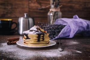 Stack of delicious pancakes with chocolate, honey, slices of banana and castor sugar on plate on wooden background, cinnamon sticks and bunch of lavender. photo