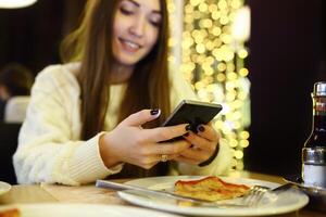 Woman typing write message on smart phone in a modern cafe. Cropped image of young pretty girl sitting at a table with pizza using mobile phone. Toned. Selective focus. photo