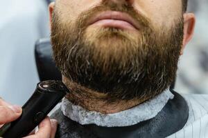 Barber trimming bearded man with shaving machine in barbershop. Hairstyling process. Close-up of a Hairstylist cutting the beard of a bearded male. photo