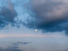 The moon peeks through a tapestry of clouds during dusk, casting a calming glow across the sky. Subtle shades of blue and gray blend harmoniously, evoking a sense of calm and introspection. photo