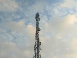 A tall telecommunications tower stands against a soft blue sky with scattered clouds, symbolizing modern connectivity and technological advancement in urban environments. photo