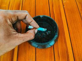 Close-up of a hand holding a cigarette above a black ashtray on a polished wooden surface. photo