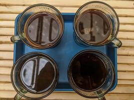 A top view of four glass mugs filled with dark coffee placed symmetrically on a blue tray atop a rustic bamboo surface. The natural lighting highlights reflections on the coffee surface. photo