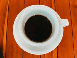 A top-down view of a white coffee cup filled with black coffee on a wooden table. The coffee is dark and strong, and the cup is the only object in the frame. photo