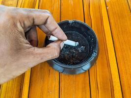 A person hand extinguishes a cigarette in a black ashtray. The wooden table contrasts with the ashtray, creating a vivid scene that represents the end of a smoking moment photo