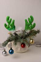 An alarm clock, a hoop with deer antlers and branches of a Christmas tree decorated with toys are placed in a cute white cup in the shape of a sleeping cat's head photo