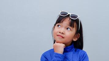 Beautiful Asian little girl wearing sunglasses thinking concentrated about doubt with hand on chin and looking up wondering photo