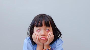 portrait of little asian girl tired, lethargic, strain, bored, bad mood concept. studio shot with light color background. pulling funny faces using their hands to pull down on their cheeks distorting photo