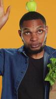 Vertical Playful vegan guy dancing with an apple placed on his head, having fun trying to be balanced and not drop it. Confident happy man recommending homegrown local fresh produce. Camera A. video