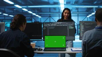 Supervisor in server hub managing diverse team of developers using green screen laptop looking for misconfigurations. Manager coordinating experts mending data center bugs with chroma key notebook video
