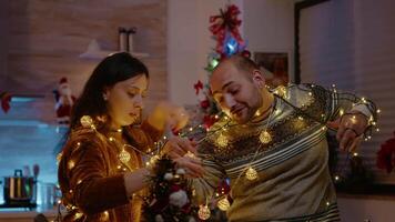 Festive couple knotted in string of christmas lights. Man and woman laughing while trying to untangle garland with twinkle lights and illuminated bulbs for holiday celebration. video