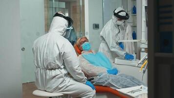 Dentist doctor in coverall showing to senior patient x-ray on stomatological monitor during coronavirus. Concept of new normal dentist visit in coronavirus outbreak wearing protective suit and face shield video