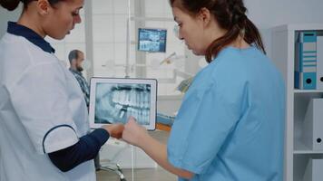 Dentist and nurse analyzing teeth radiography on tablet for modern oral care examination at dentistry clinic. Professional stomatological team using digital device for patient teethcare video