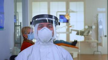 Portrait of exhausted doctor in dental office looking on camera wearing ppe suit and face shield sitting on chair in waiting room clinic. Concept of new normal dentist visit in coronavirus outbreak. video