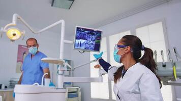 Patient pov to dentist talking about mouth hygine showing x-ray on monitor. Stomatology specialist wearing protective face mask and gloves pointing at teeth radiography in stomatological clinic video