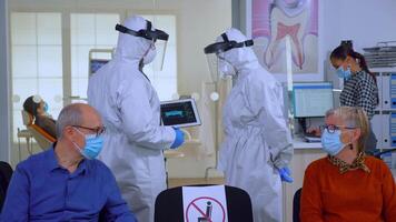 Man discussing with nurse in dental reception wearing protection suit against coronavirus, elderly patients waiting in reception keeping distance. Concept of new normal dentist visit in outbreak. video