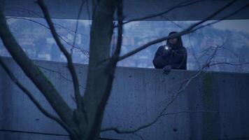 A man standing alone by a concrete wall under a bare tree in the bluetoned evening video